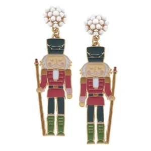 Pair of enamel earrings- each earring is a 2.5" tall nutcracker wearing a red coat, black pants, knee high green boots, and a black hat. One hand on each nutcracker holds a wood staff. The nutcracker sports the traditional white beard down his chest. The nutcrackers both hang from the top of their hats on pearl-colored studded earring posts.
