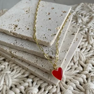 A gold paperclip chain necklace drapes across a stack of cream colored stone coasters to display the a dainty red enamel heart trimmed in gold.