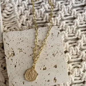 A delicate gold chain necklace is draped across a white stone background featuring a replica St Benedict aged gold coin.