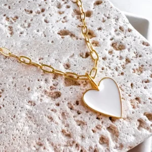 On a cream color stone background, the Heart on My Sleeve 18" Necklace in White lays featuring the gold paperclip chain and the white enamel heart on gold fill.