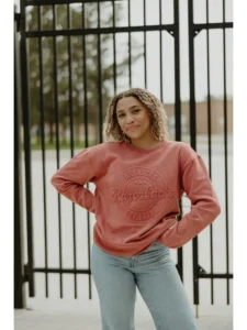 A young lady stands outside with her hands on her hips. She is wearing a cardinal red sweatshirt featuring a reverse-embossed Arkansas Razorbacks Est. 1861